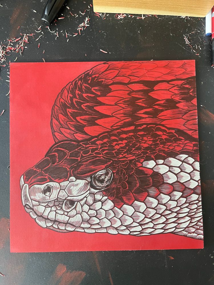 Red linoleum block with a rattlesnake drawn on it. Some of the scales on the face have been carved.