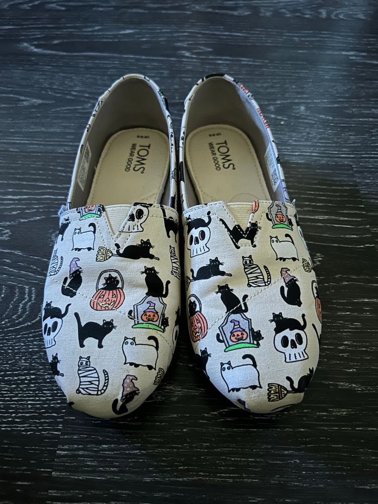 Close up of a pair of Tom’s canvas shoes. The shoes are covered in a cartoon Halloween print with black cat mummies, black cats in pumpkins, black cats on top of skulls,etc.
