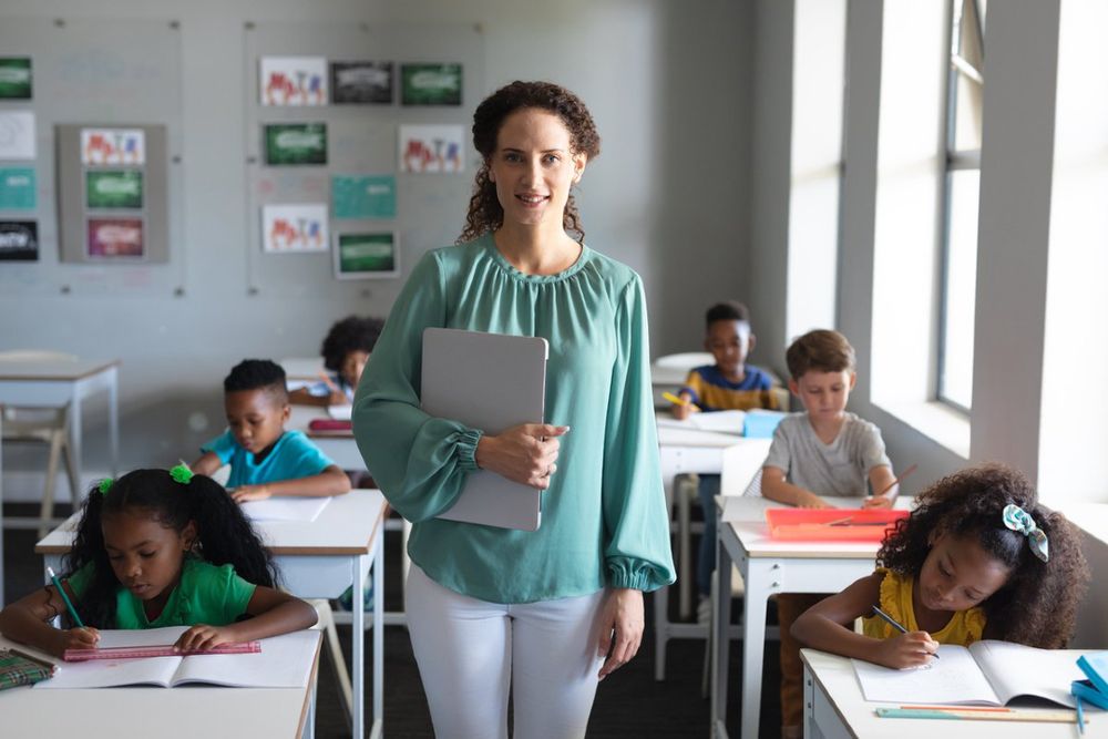 A teacher holding a laptop stands at the front of a classroom with diverse students focused on their work at desks.