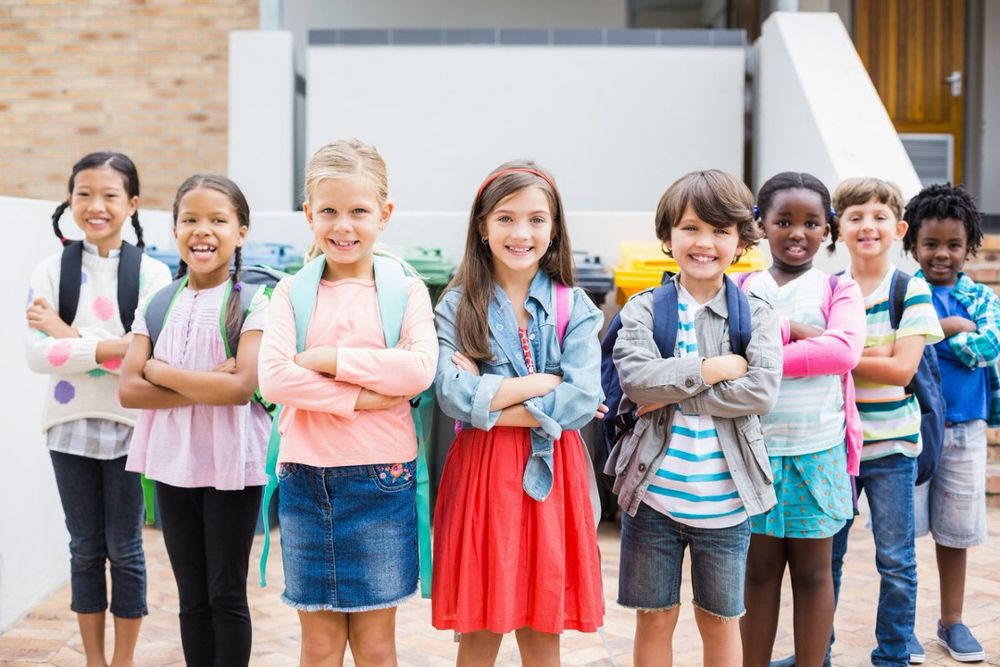 A diverse group of smiling children with backpacks stand confidently in two rows, posing in front of a school building.