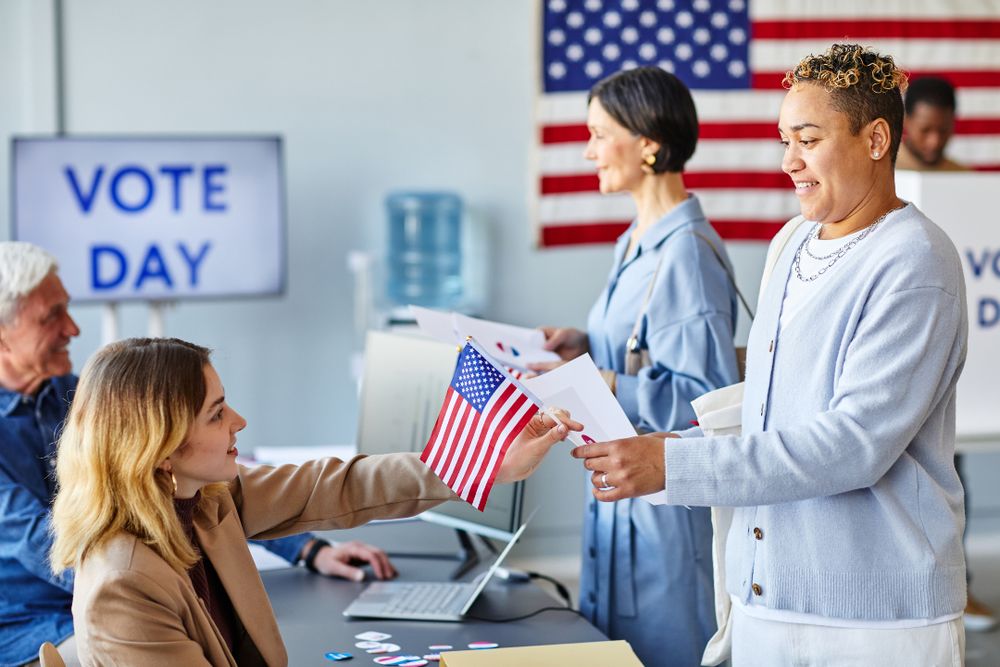 A smiling voter hands an American flag to a polling station worker, with "Vote Day" displayed in the background on a screen.
