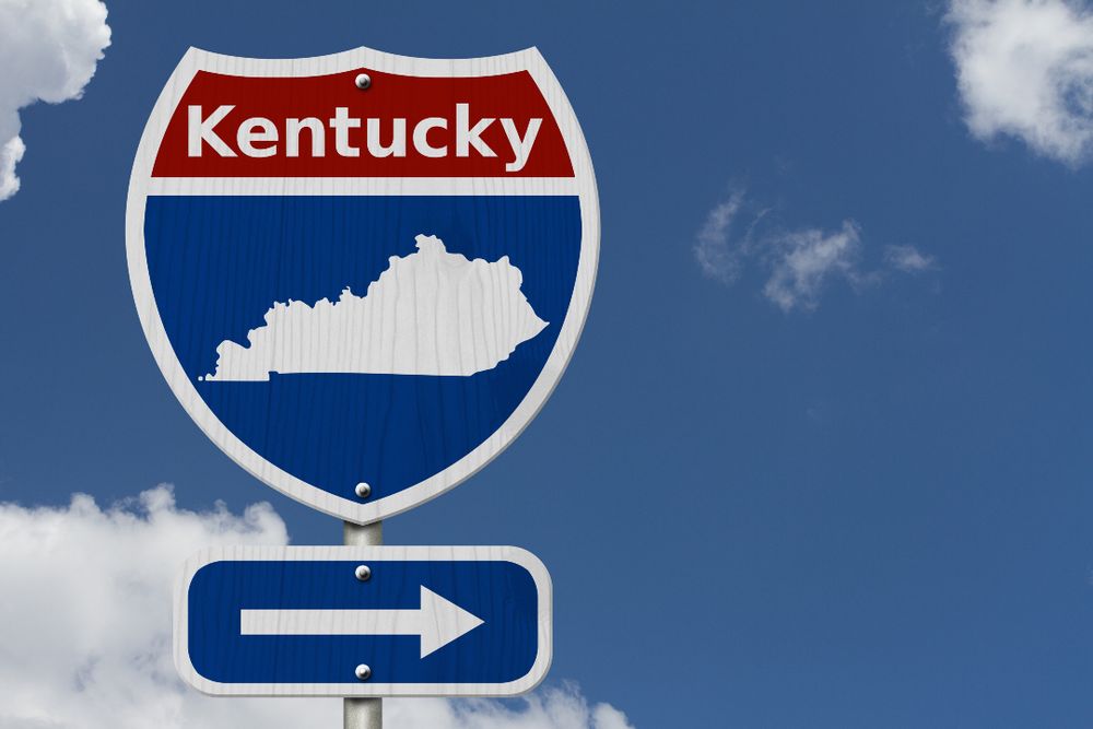 Highway sign with the word "Kentucky" above a silhouette of the state and a directional arrow pointing right against a blue sky.
