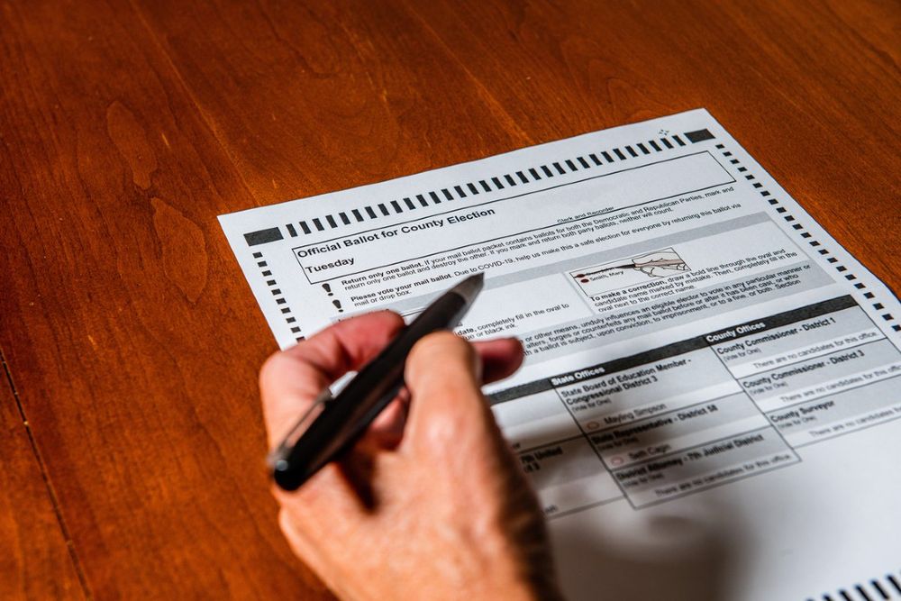 A hand marks an official county election ballot with a pen, focusing on filling in choices for various positions and measures.