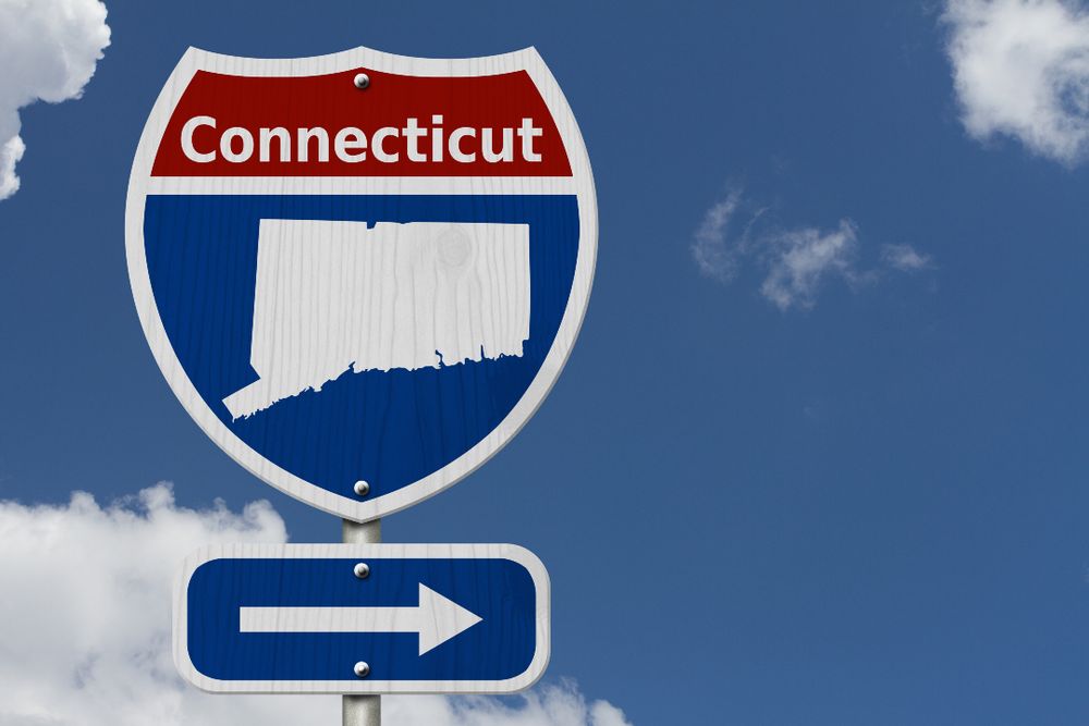 Highway sign with "Connecticut" and the state's silhouette, above a blue arrow pointing right, set against a partly cloudy sky.