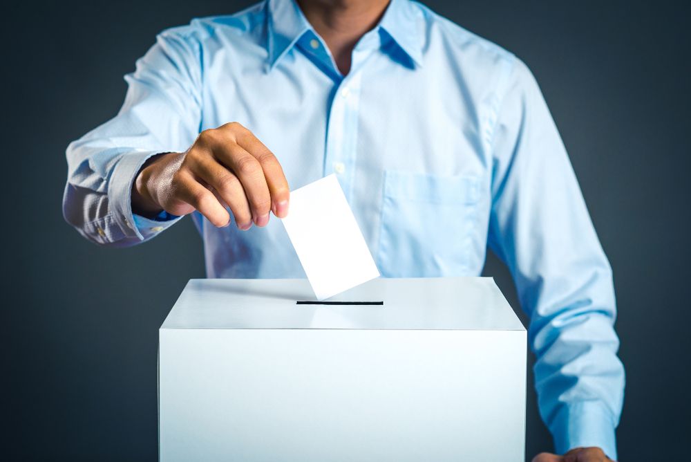 Person in a light blue shirt casting a vote by placing a ballot into a white box against a dark background.