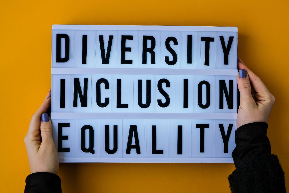 Hands holding a board with the words "Diversity Inclusion Equality" in bold black letters on a bright orange background.