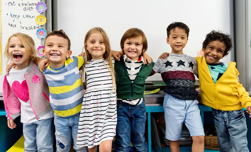 Six diverse children standing in a classroom, smiling and with arms around each other. Classroom rules are partially visible on the left.