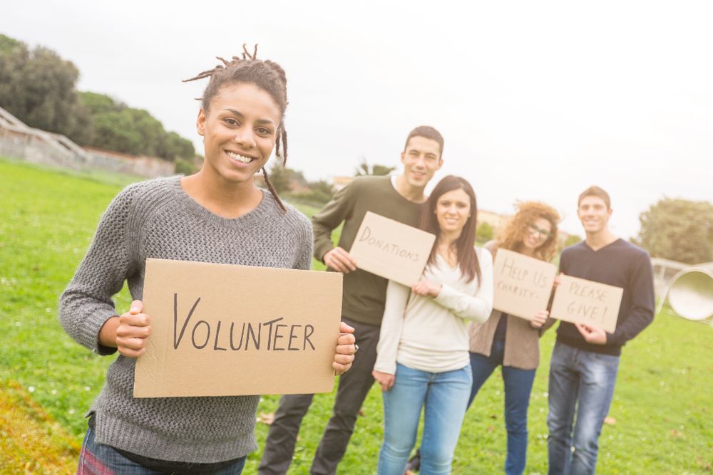 Group of young adults holding signs encouraging volunteerism and donations, smiling in a park setting. The focus is on a woman holding a "VOLUNTEER" sign.
