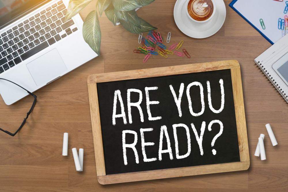 A chalkboard with "ARE YOU READY?" on a desk, surrounded by a laptop, coffee, paperclips, a plant, and a notebook.