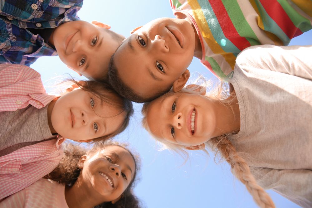 Five smiling children from diverse backgrounds huddle together, looking down at the camera against a clear blue sky.
