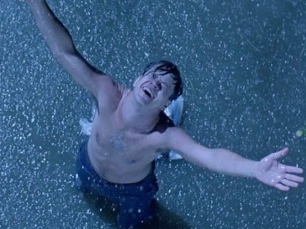 A man stands looking upward with arms outstretched and shirt off in the rain 