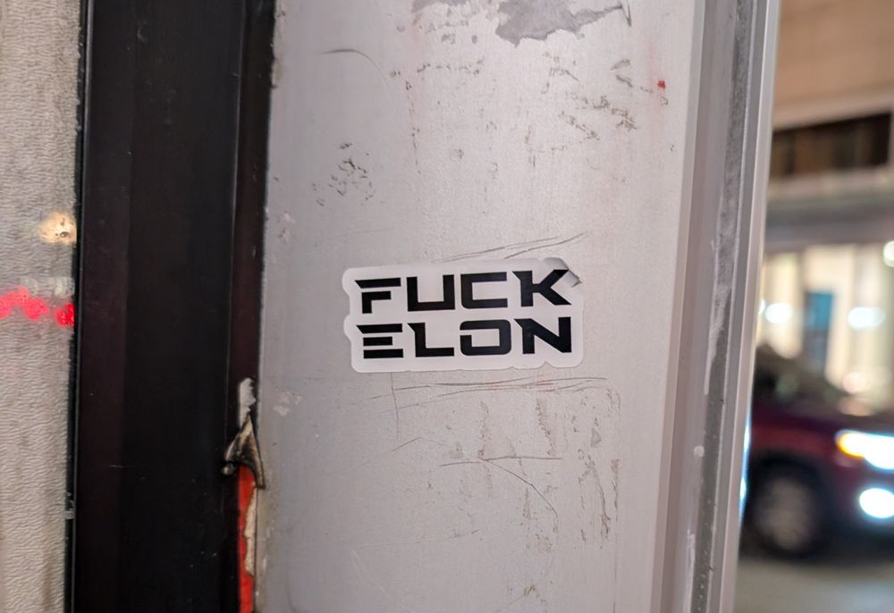 A sticker on a public structure that reads "FUCK ELON" in a font similar to Tesla's branding