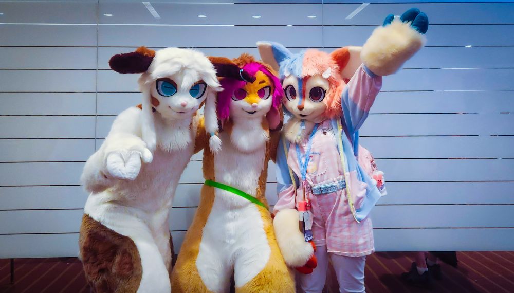 Three LikeaPalette fursuiters taking a group photo. From left to right, Fonn a white a brown deer, Kyoho a tan and purple Rabbit, and HoneyxFawn a blue and pink deer.