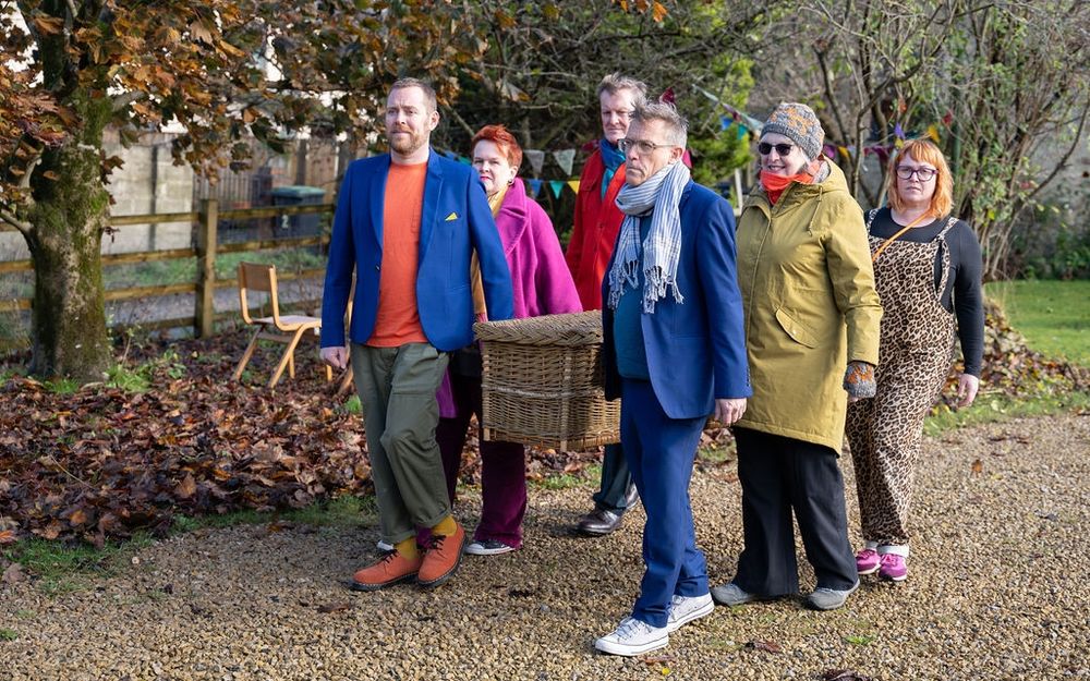 Six adult friends in bright clothing carrying a locally made willow coffin at waist height along a garden path