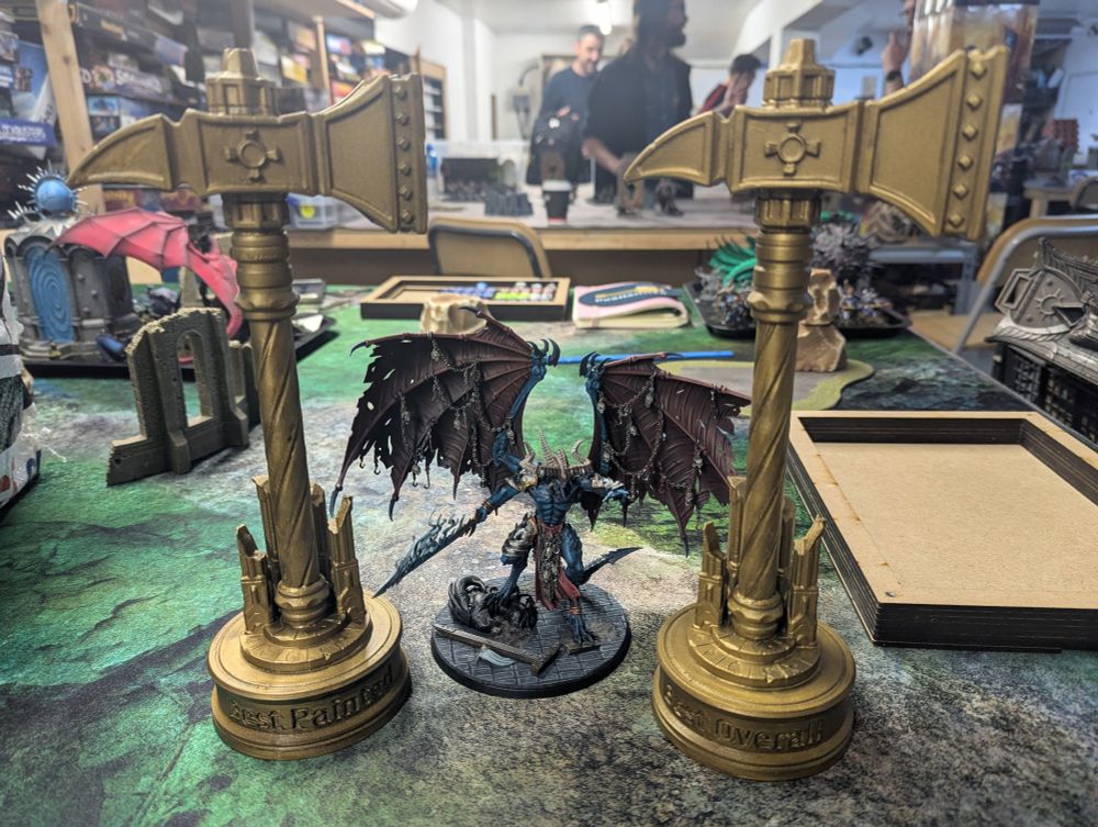 Two gold warhammer shaped trophies, one labelled best painted and one labelled best overall. The Be'lakor model from Games Workshop is standing between them.