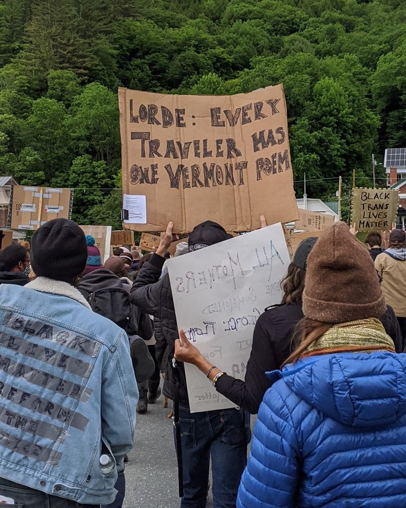 Protesters at a Black Lives Matter march. The primary sign in the frame says, "Lorde: Every Traveler Has One Vermont Poem".