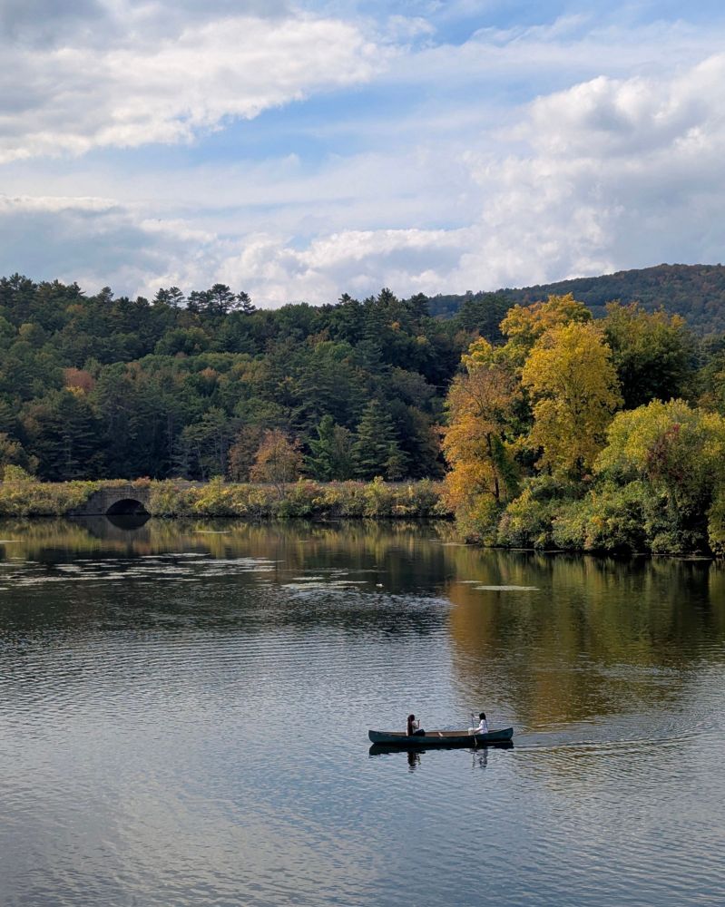 Two people sitting in a canoe on the Connecticut river, facing each other. Behind them, a small bridge supporting train tracks, trees beginning to turn orange and yellow. In the distance, a hill, and a partially cloudy sky.