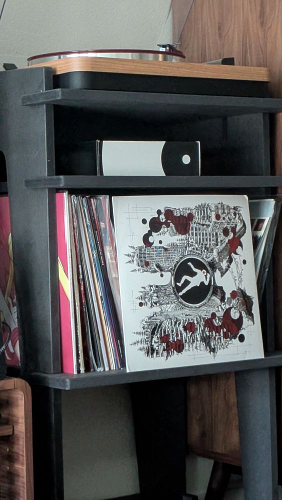 A record player on top of a record shelf with a record out front. The record is white, black, and red with a black circle in the middle. In the black circle there is a white cutout of a body, possibly dead. It "goes hard" as the kids say. 