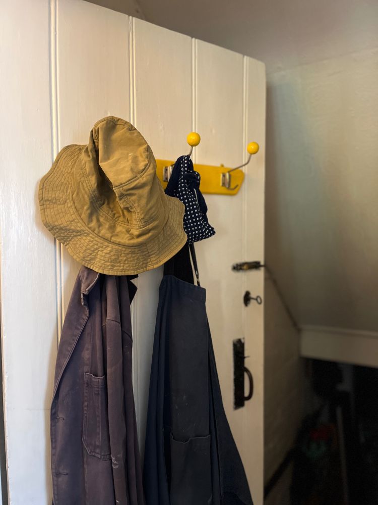 A white wooden door is open to a dark basement. On the door is a yellow handpainted coat rack. On the lefthand hook is a man’s khaki gardening hat, and a faded blue workjacket. On the centre hook, beside it, is a woman’s blue-and-white-spotted headscarf and a navy apron. Portrait of a long marriage as workwear.