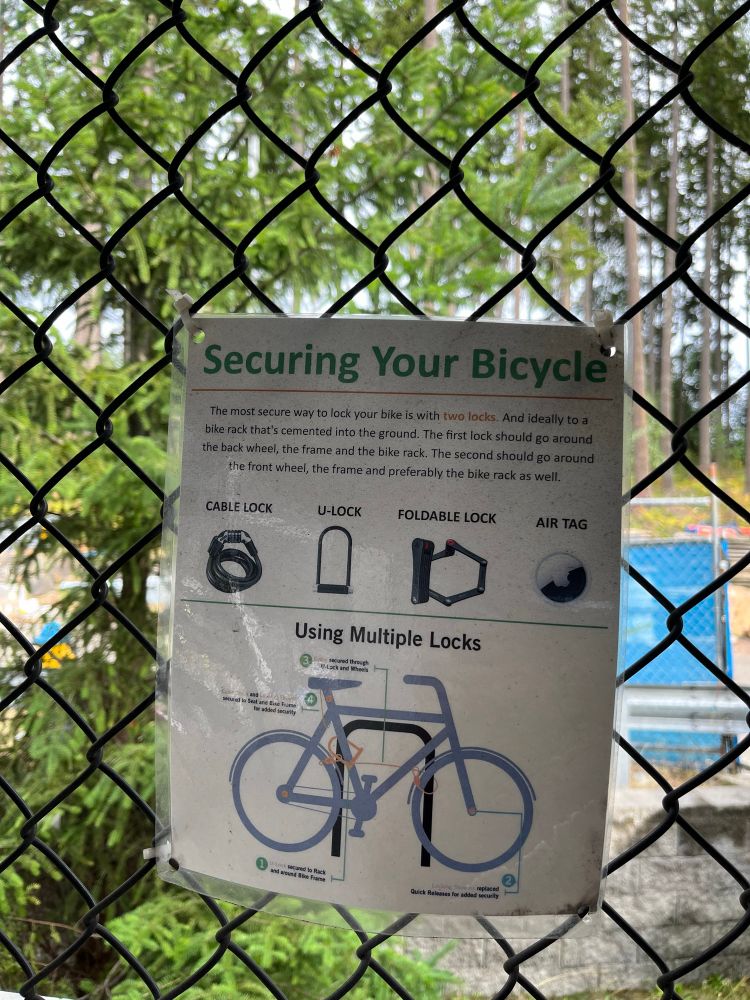 A letter sized sign secured to a fence above some grid bike parking. Shows an image of a bicycle locked with a chain and a u lock to a staple. 