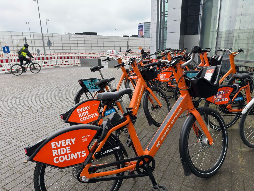 Bunch of orange rental bikes with signs saying “Every ride counts.” Cyclist in background.  Airport terminal location.  