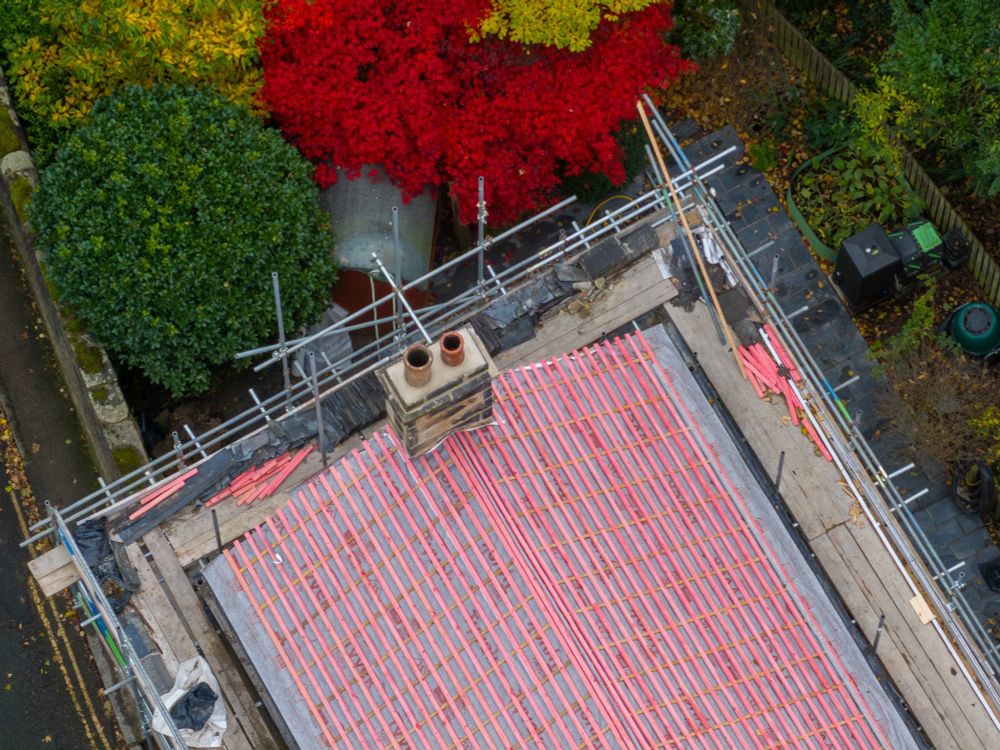 House with red showing through tiles and red tree in amongst green and yellow trees next to cottage. Autumn colours. Close drone shot.