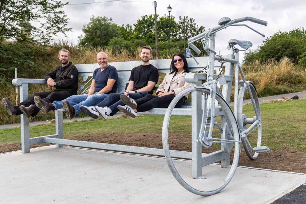 Four people sit on oversize metal bench with legs up next to oversize bicycle locked to bench.