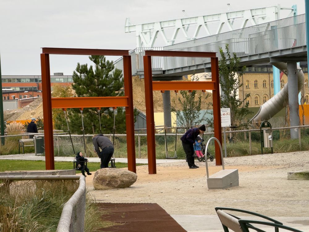 Two toddlers pushed in swings by fathers. Swings are suspended from reclaimed girders. 