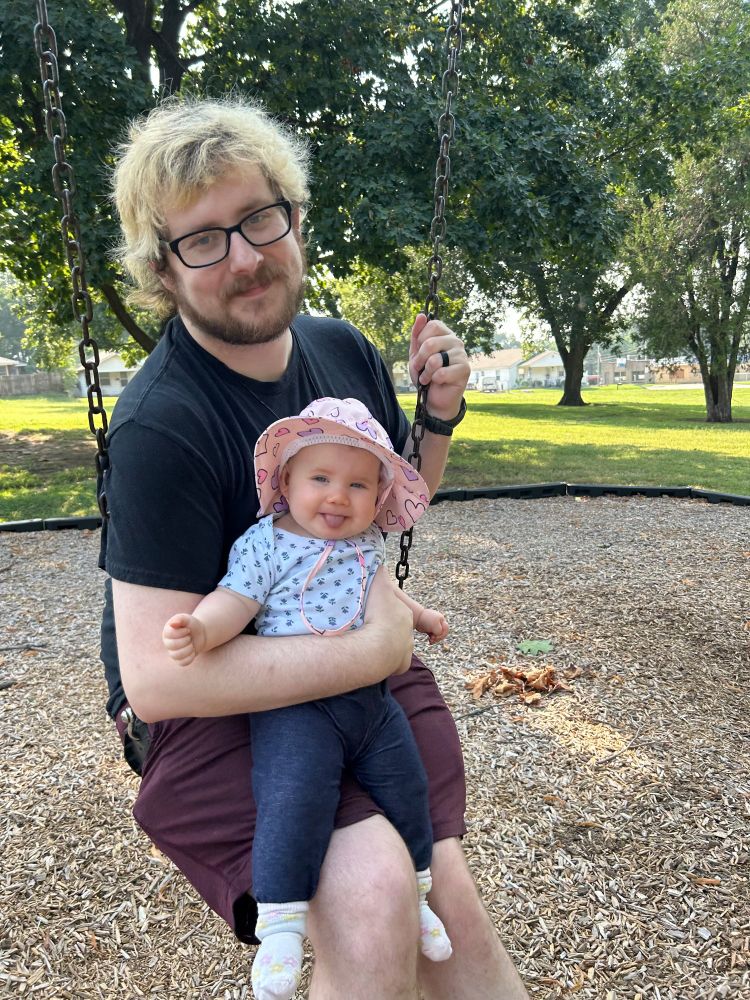 a guy on a swing in a park holding a small baby, who is being adorable with her tongue out and wearing a sun hat