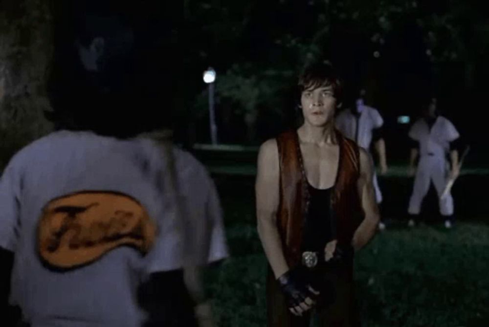 a baseball player with a mask on his face is holding a bat and talking to another man .