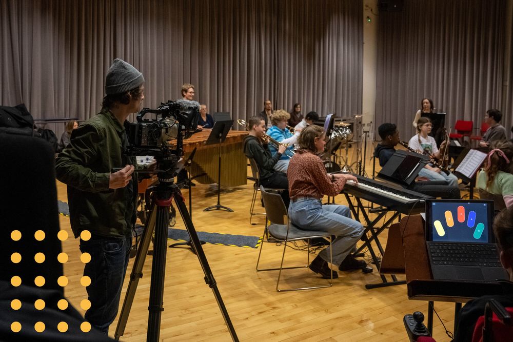 A camera operator films an orchestra rehearsing. The orchestra members are disabled and non-disabled and play a mix of acoustic and electronic instruments.