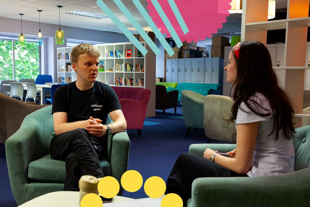 Torin sits in the BSO offices talking to Hannah, a member of the BSO team. They are sitting on green armchairs. Torin wears a black t-shirt with white writing on it and dark trousers. Hannah wears a white pin-striped shirt and dark trousers.