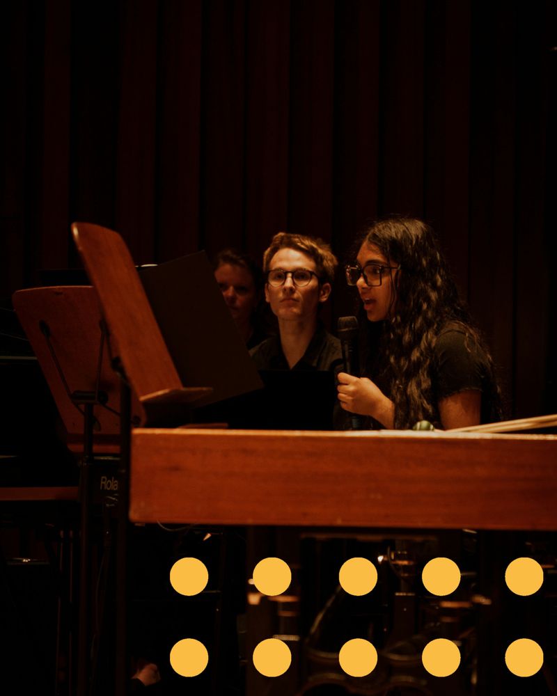 A young female musician sits behind an electric piano on stage and talks into a microphone.