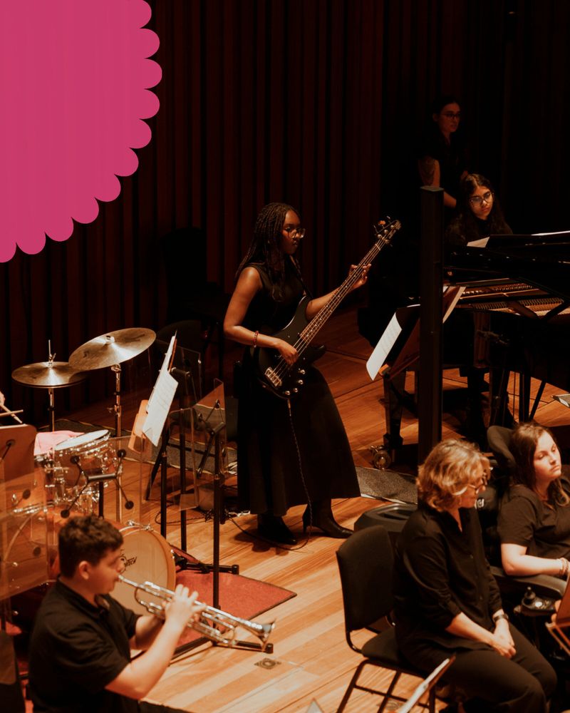 A young female musician plays bass guitar on stage. She is surrounded by other members of the National NOYO Ensemble.