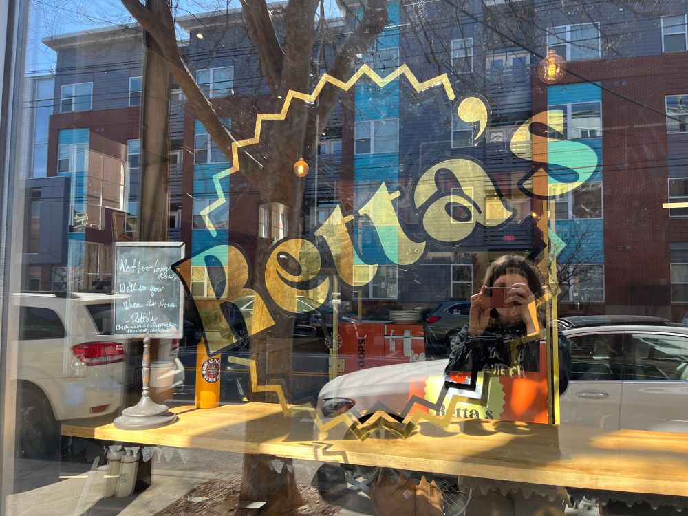 A window with gold leaf and a painted sign with a reflection of buildings and a person in the glass