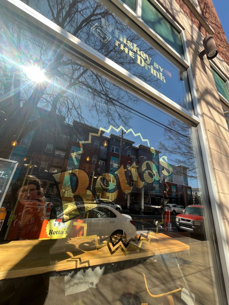 A window with gold leaf and a painted sign with a reflection of buildings and a person in the glass