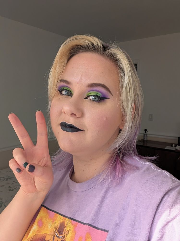 Another selfie of me, with different bold makeup, this time in lime green and purple, with black lipstick. I'm smiling and giving a peace sign.