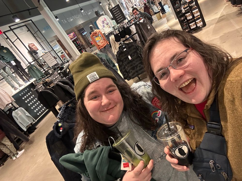 Two women smiling at the camera while holding coffee in a clothes store.