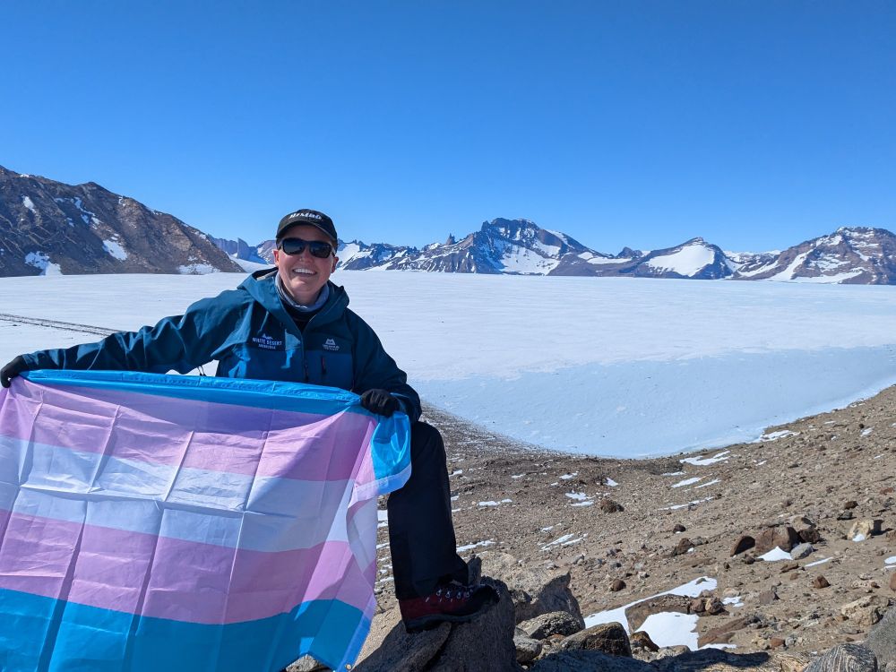 Sophie stands on a barren ridge in Antarctica, holding a trans flag. A glacier and snow-covered mountains lie in the background. 