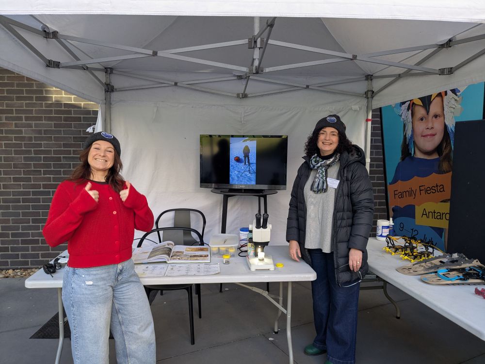 Scientists Madi and Bec in front of a table with microscopes, snowshoes and crampons. 