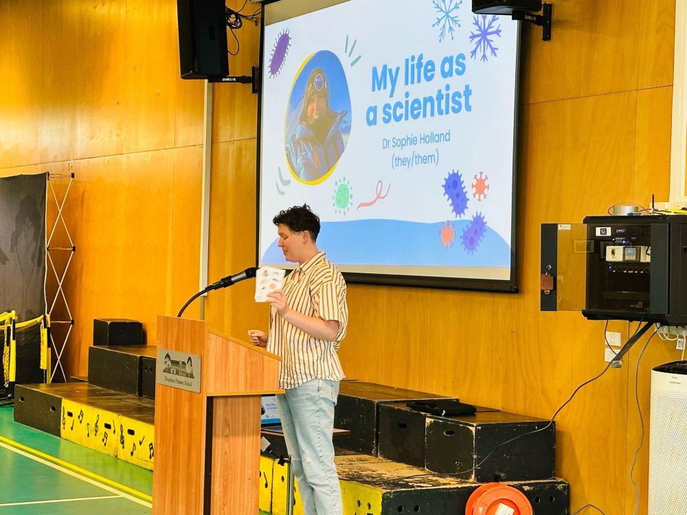 A photo of Sophie behind a podium, in front of a projector screen with their title slide "My life as a scientist".