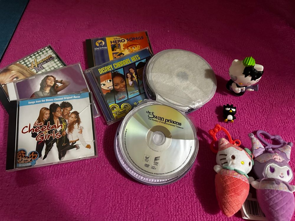 Image shows multiple CDs, some of which are Hannah Montana, Miley Cyrus and the cheetah girls. Also the Swan, Princess DVD, a Disney Channel hits CD, and a Disney’s heroes CD. Also a hello Kitty ice cream plushy and a Kurumi plushy.