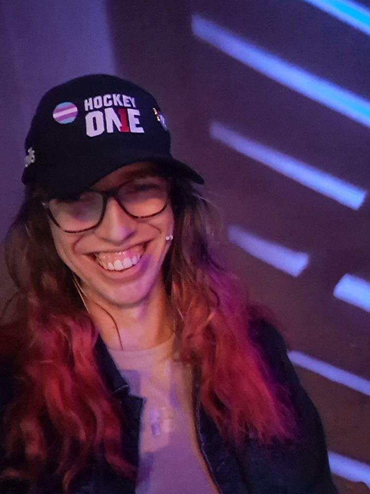A white woman smiling at the camera wearin a jean jacket and a baseball cap. The cap says "hockey one" on it and has a trans flag pin. She's wearing glasses and a headset mic, and has dyed red hair.