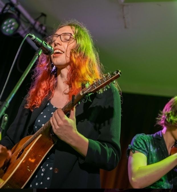 Zoe singing into a microphone with her eyes closed, wearing a polka dot dress and playing an octave mandolin, in green and purple lighting