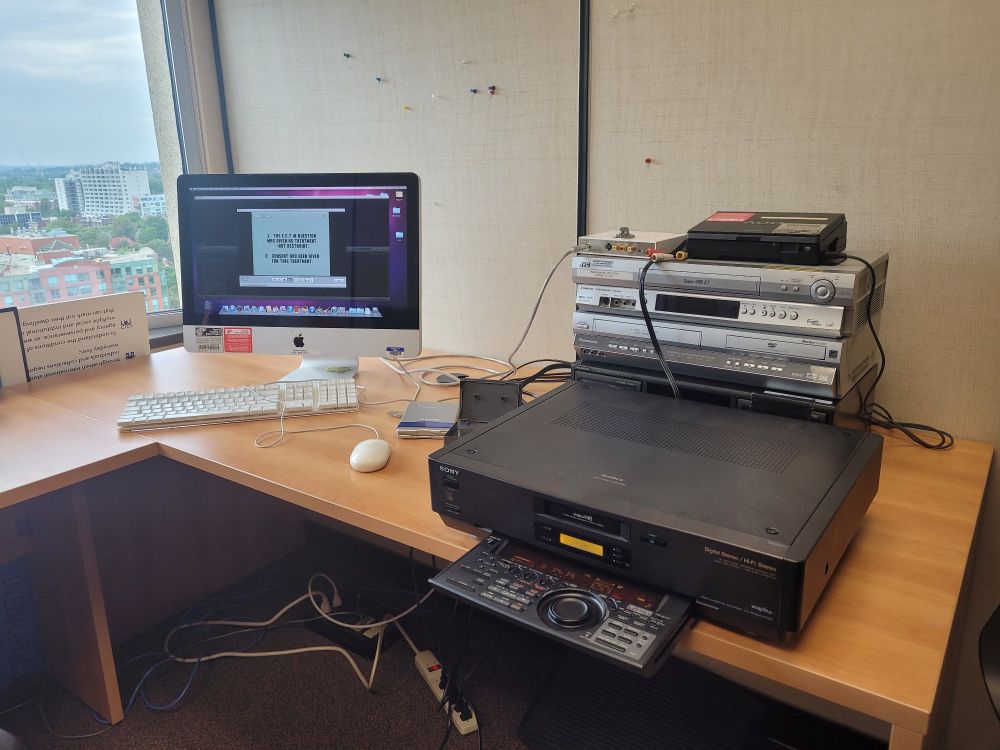 On the left, a computer screen. On the right, a stacked pile of old technology including a hi-8 player, cassette player, VHS/DVD player, and HDV player