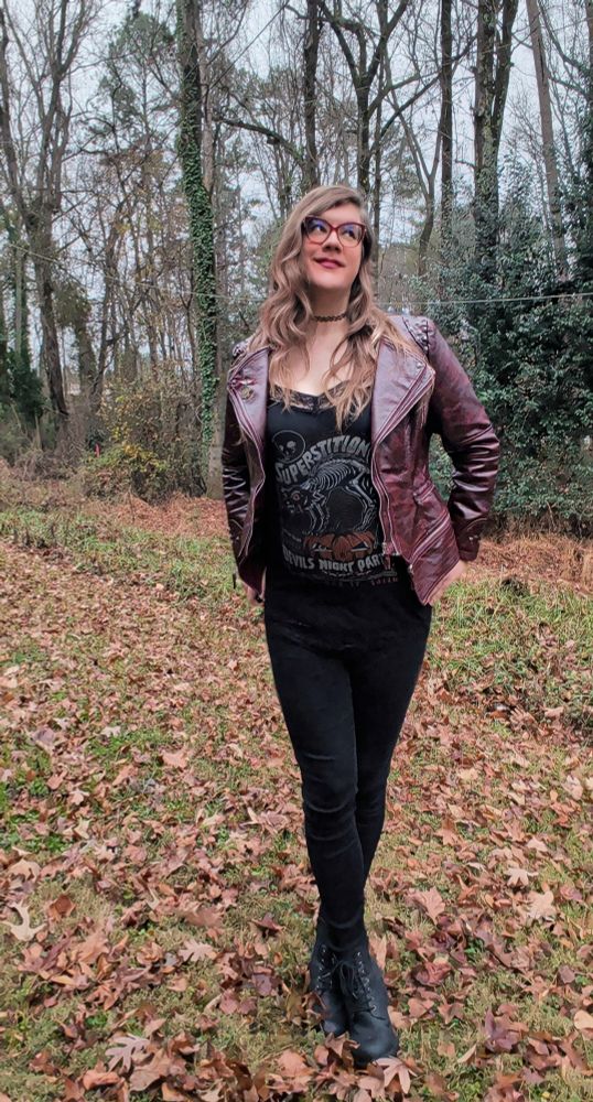 A picture of Robyn outdoors, amongst the leaves and trees. She's wearing a black top, black leggings and boots. She's also wearing a red leather jacket 