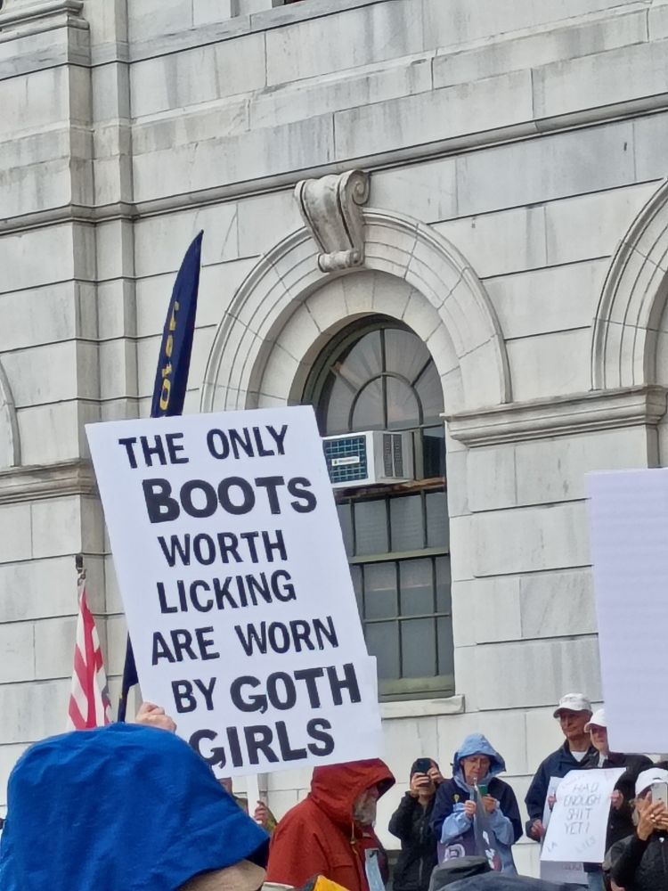 A protestor holding up a sign that reads 
"The only boots worth licking are worn by goth girls"