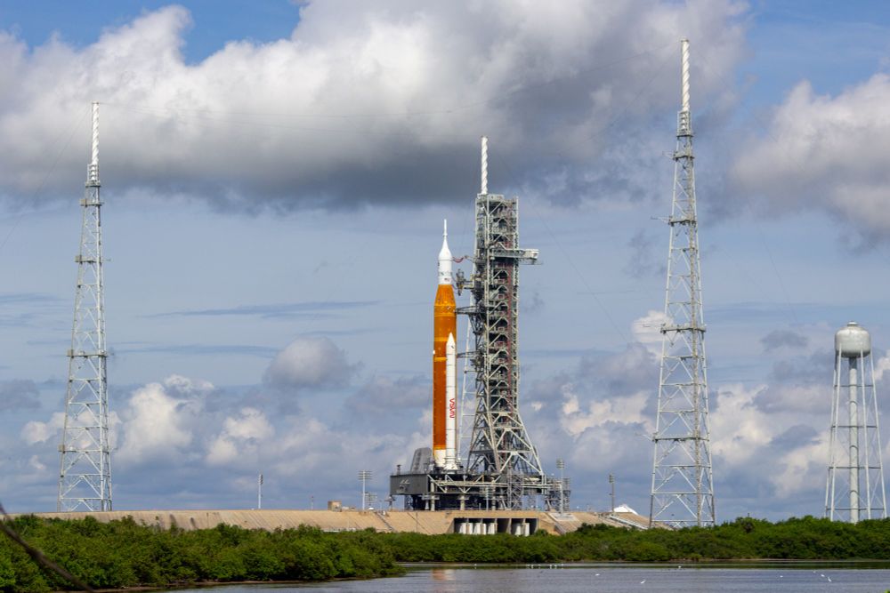 NASA's Space Launch System rocket is on its launch pad, with the large Mobile Launcher Tower next to it. The rocket has two lighting towers either side of it, with a water tower on the far right of the frame.

Below the rocket is the concrete and brick flame trench, with a small swampy area in the foreground.