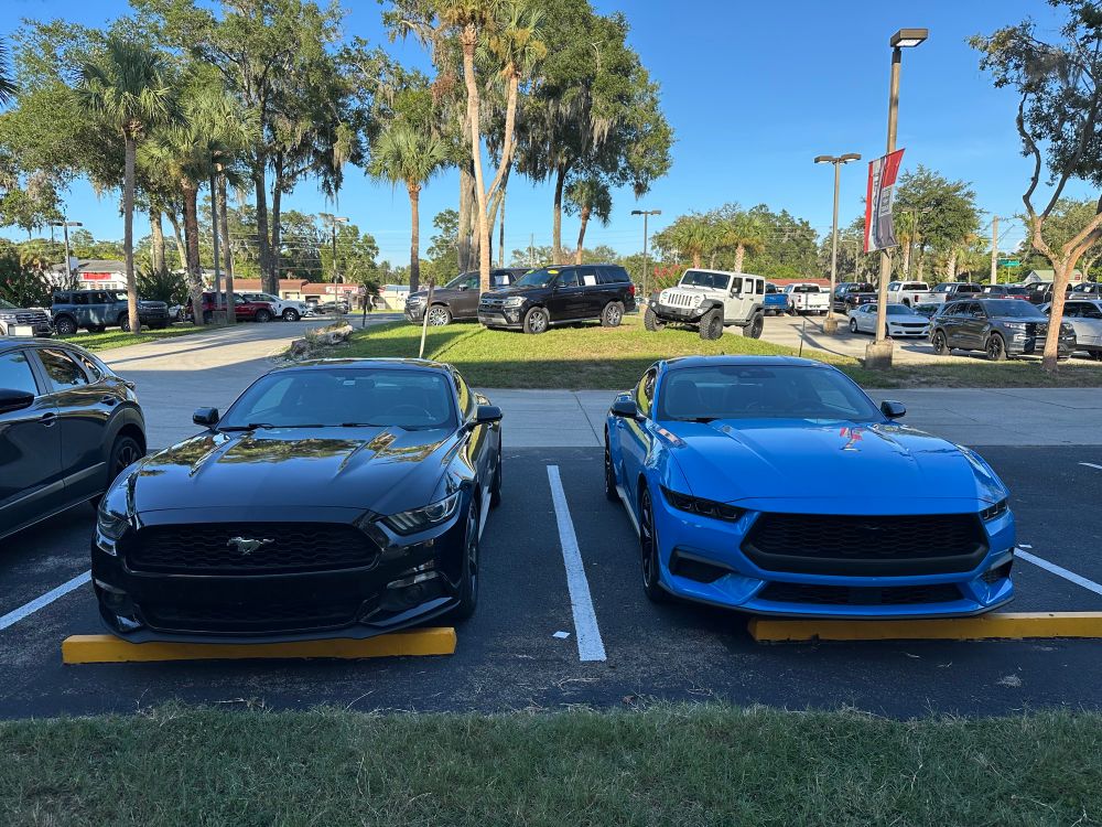 A pair of Ford Mustangs sitting next to each other. On the left is a 2015 in Black, on the right is a 2025 in Grabber Blue with a black top and black mirrors.