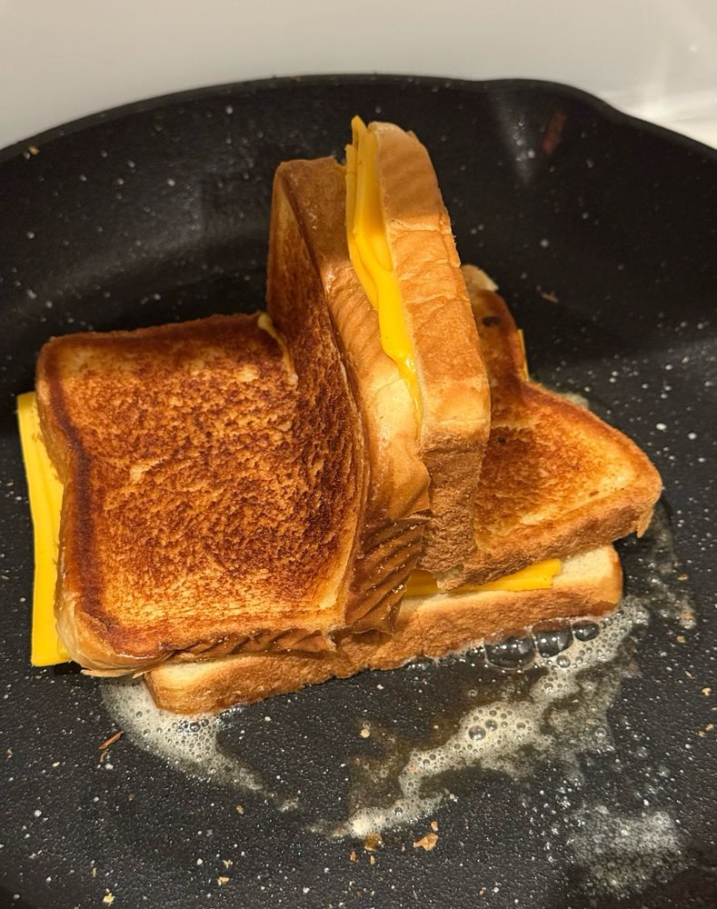 A three-sided grilled cheese made with white bread and American cheese, cooking in a cast iron pan. The visible sides of the sandwich are golden brown